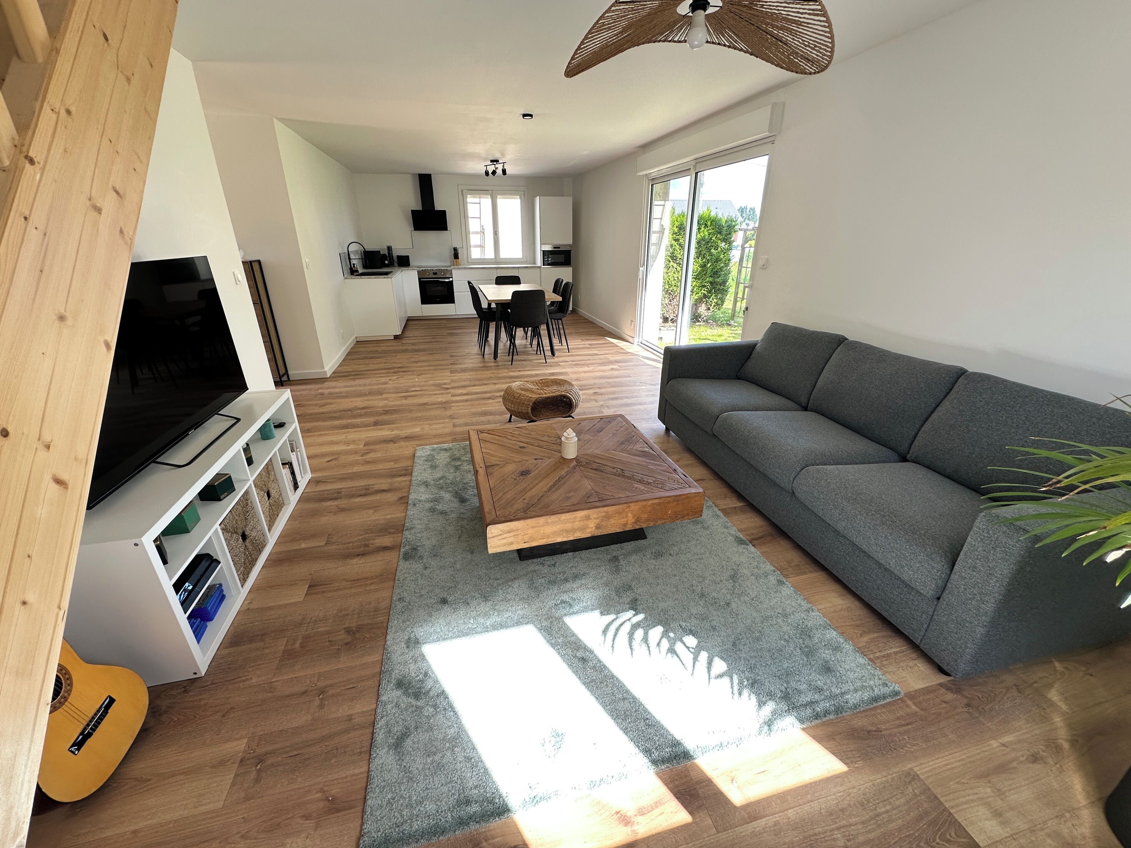 A modern living room with a flat-screen TV, a wooden coffee table, a grey sofa, and a dining area in the background.