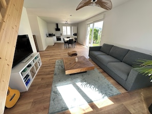 Ein modernes Wohnzimmer mit Flachbildfernseher, Couchtisch aus Holz, grauem Sofa und Essbereich im Hintergrund.