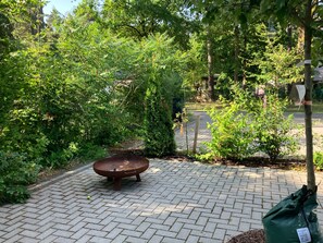 A paved outdoor area with a metal fire feature, surrounded by dense greenery and trees.