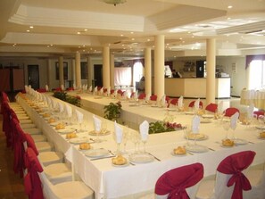 A banquet hall with long tables set for a formal event, featuring white tablecloths, red chair covers, and glassware.