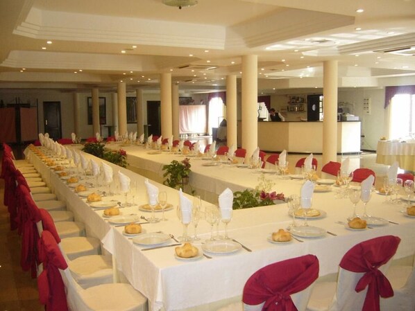 A banquet hall with long tables set for a formal event, featuring white tablecloths, red chair covers, and glassware.