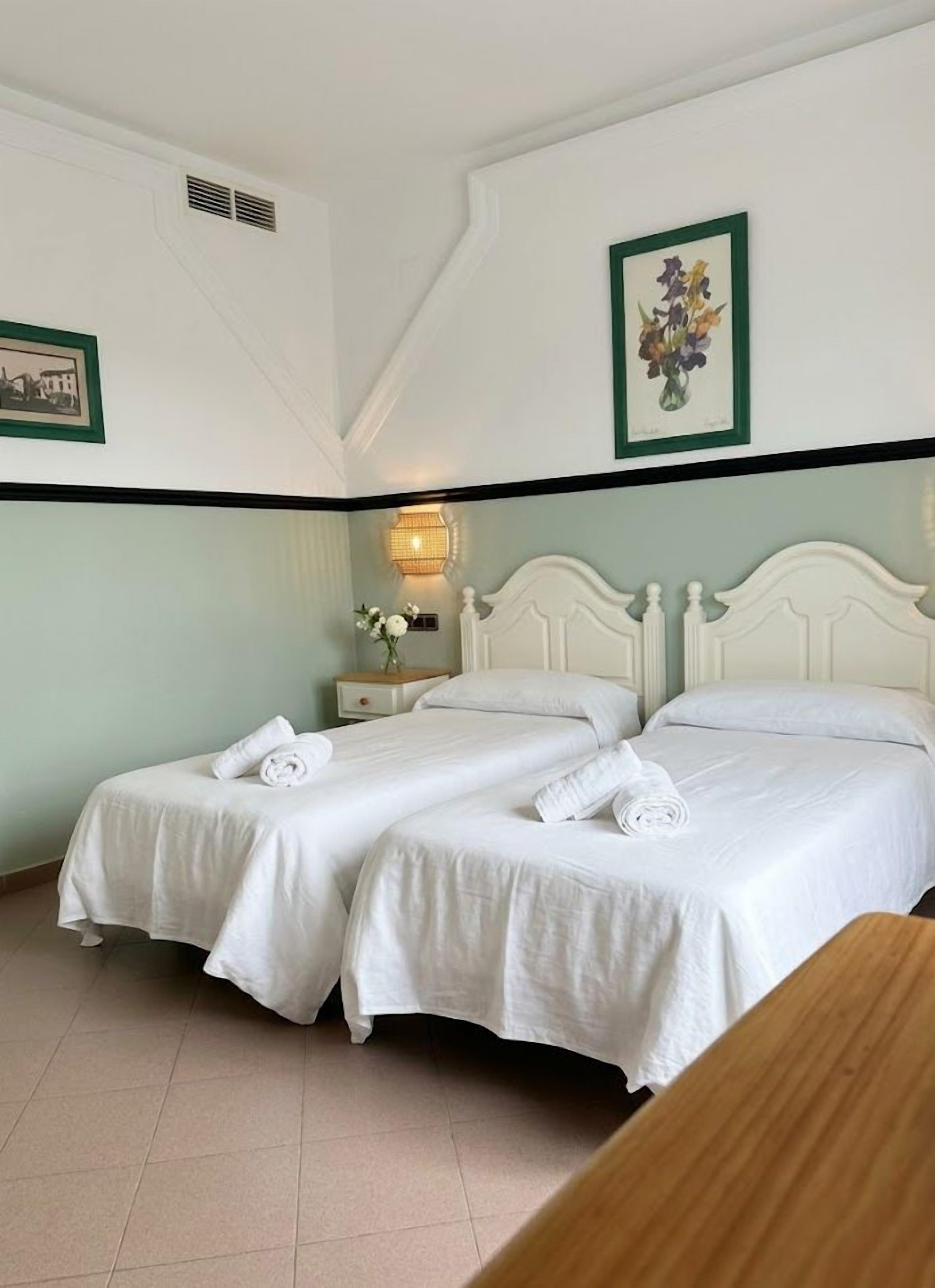 Habitación de hotel con dos camas, una mesita de noche con lámpara e impresión botánica enmarcada en la pared.