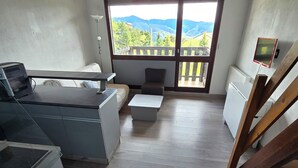 Um quarto com teto de madeira, uma janela grande com vista para montanhas, um sofá, uma mesa pequena e um radiador.