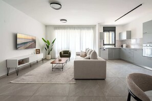 A modern living room with a sofa, a coffee table, a TV, and a plant. - Elegant 4BR Stay l Balcony & Mamad (Bat Yam)