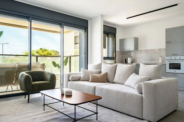 Un moderno salón con un sofá, una mesa de centro y una silla. Hay un balcón con vistas a zonas verdes.