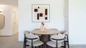 Une table de salle à manger ronde, entourée de six chaises, un vase contenant des fleurs séchées et, au mur, un tableau abstrait encadré.