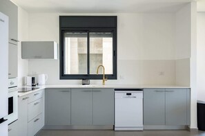 A modern kitchen with grey cabinets, a white dishwasher, a coffee maker, and a window with a black frame. - Stylish 4BR Apt l Private Balcony & Mamad (Bat Yam)