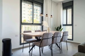 A modern dining area with a wooden table and six chairs, large windows with curtains, and hanging pendant lights. - Stylish 4BR Apt l Private Balcony & Mamad (Bat Yam)
