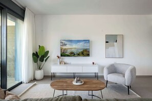 A modern living room with a large window, a flat-screen TV, a white console table, a grey armchair, and a wooden coffee table. - Stylish 4BR Apt l Private Balcony & Mamad (Bat Yam)