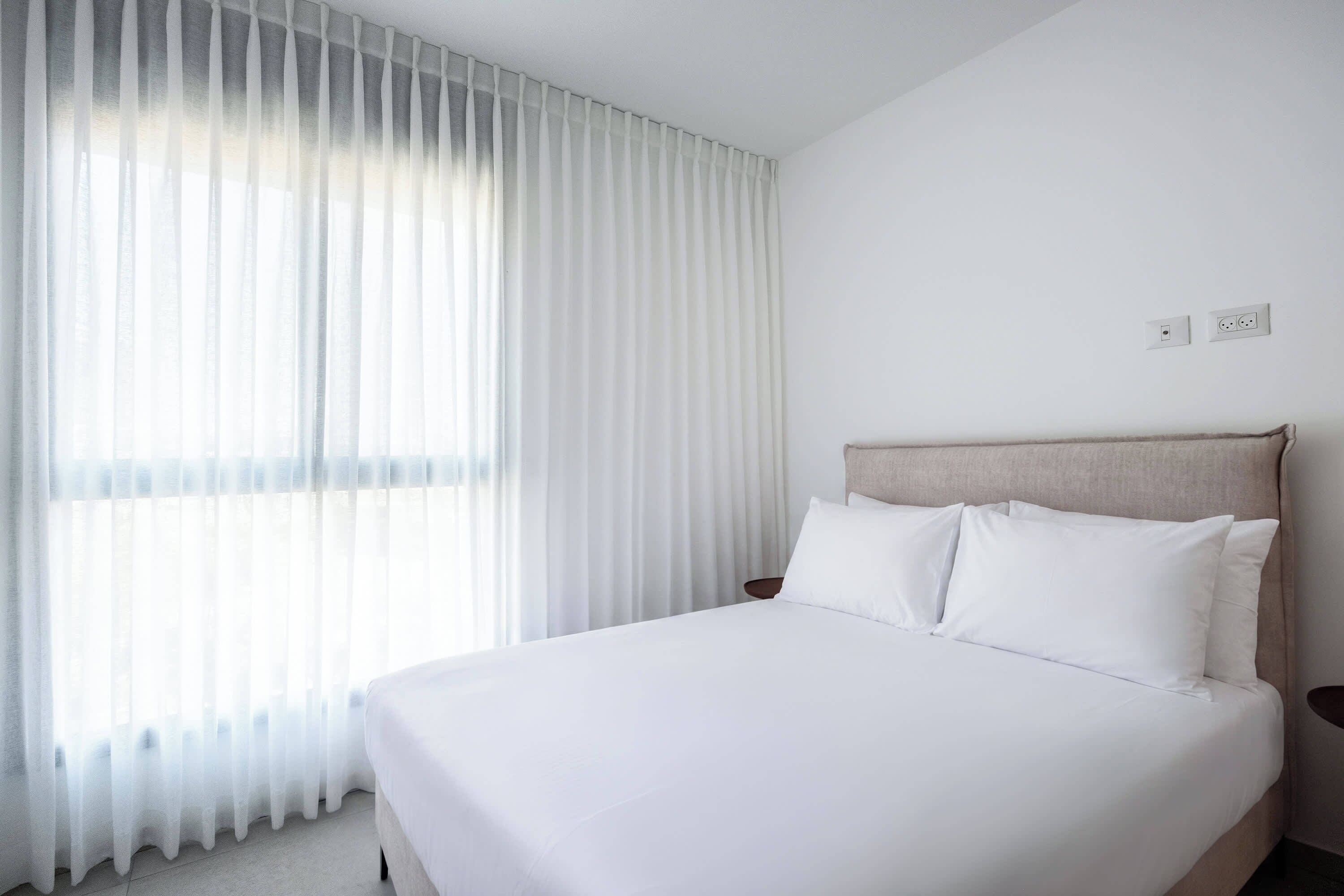 Serene bedroom with expansive sheer curtains, natural window light, beige headboard, and tranquil minimalist atmosphere.
