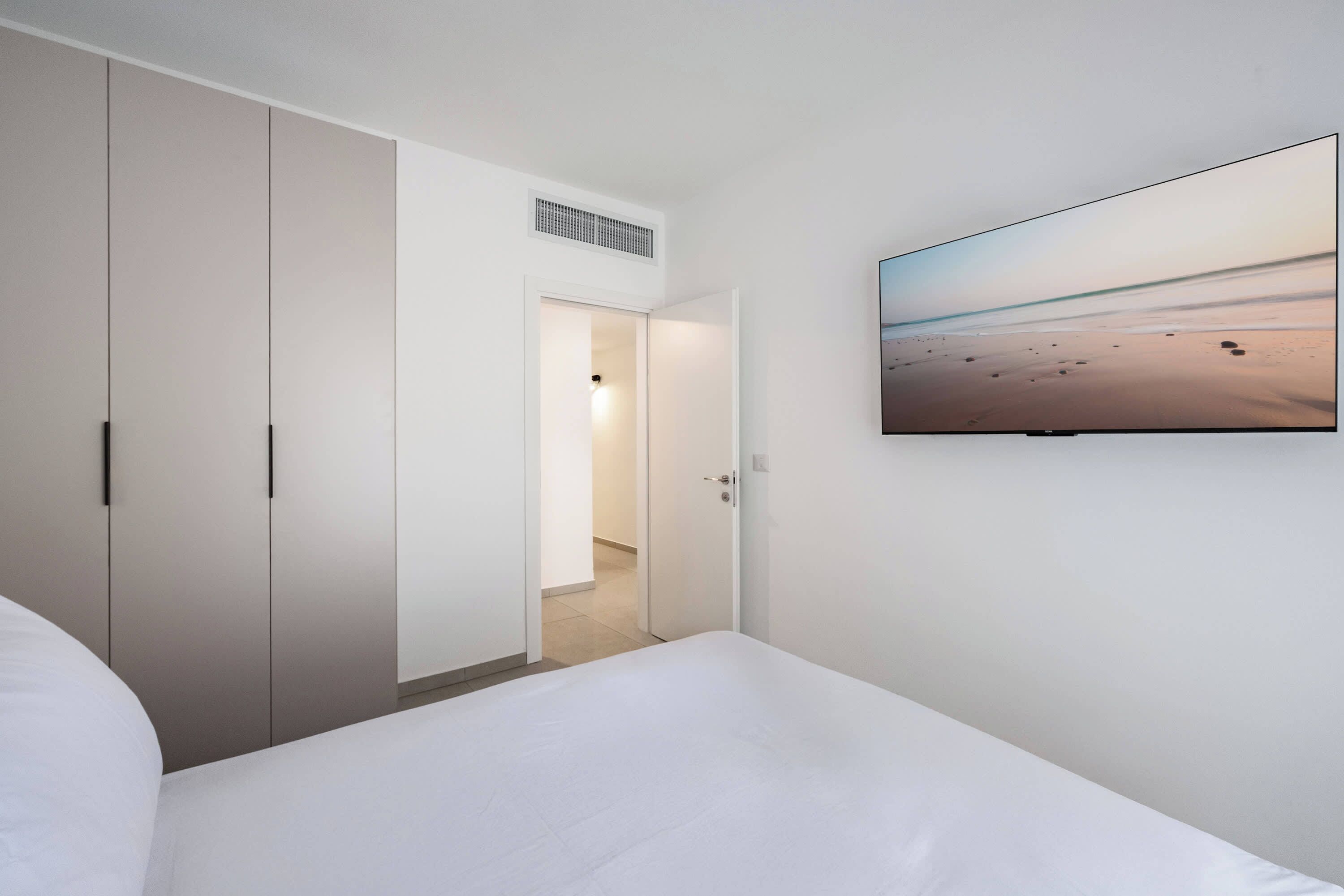 Een hotelkamer met een bed, een kledingkast, een deur naar een badkamer en een tv aan de muur waarop een strand te zien is.