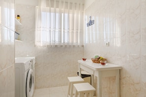 A bathroom with a washing machine, a small table with a basket of fruit, and a window with sheer curtains. - Apartment 'Ajuda Mar Holidays with Parking' with Balcony and Wi-Fi (Funchal)