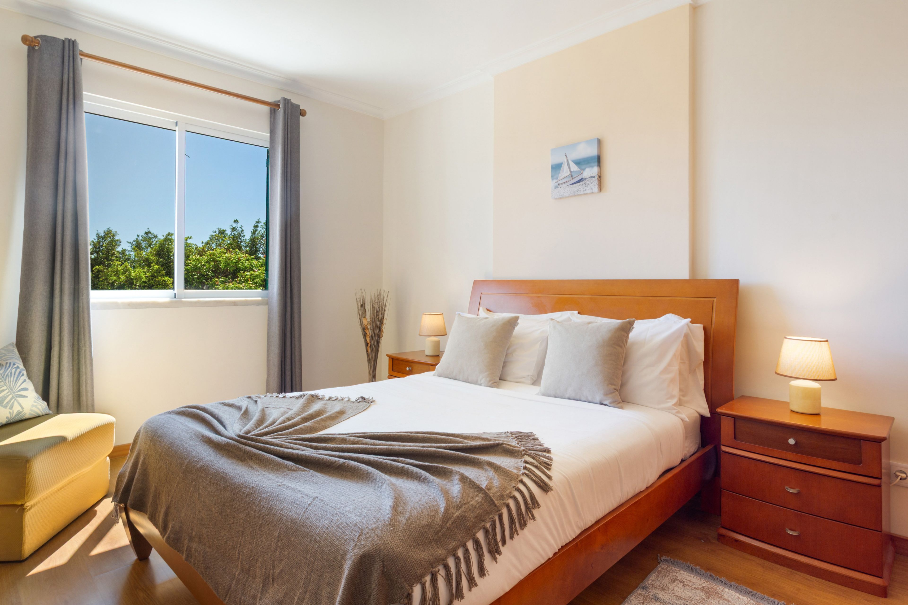 A hotel room with a bed, bedside tables, a lamp, a window with curtains, a painting on the wall, and a view of trees outside.