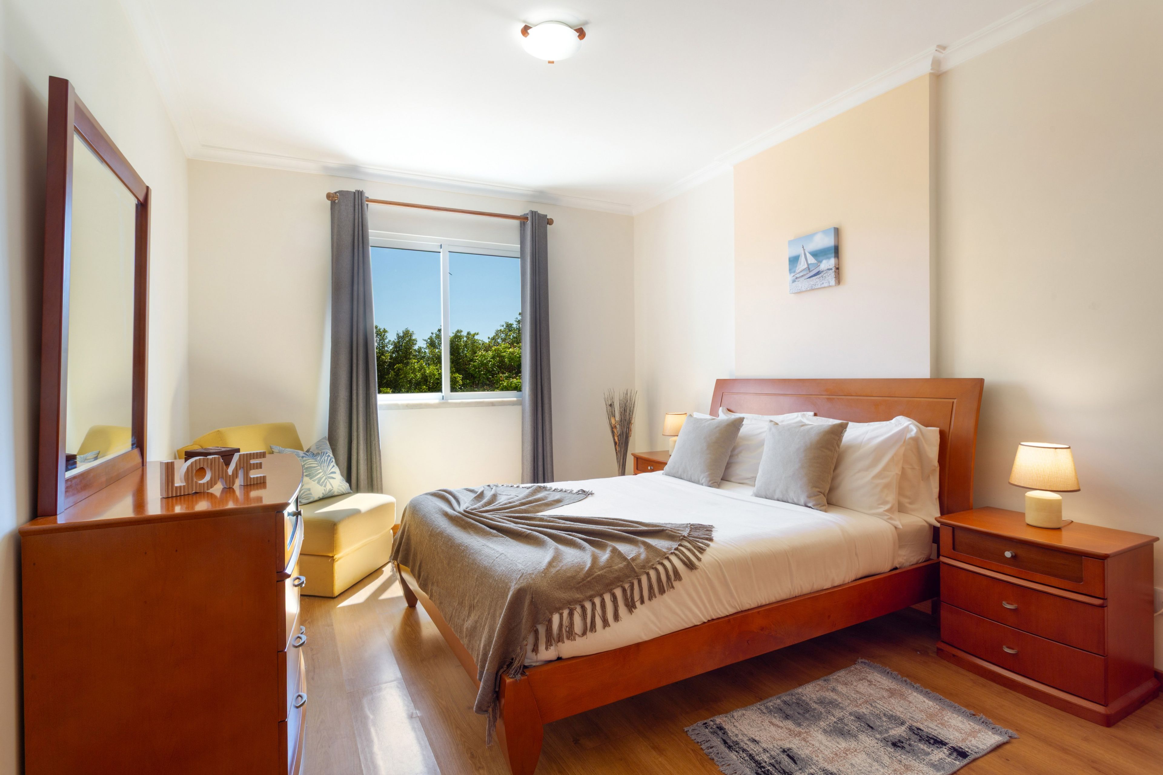 Een hotelkamer met een bed, nachtkastjes, een dressoir, een raam met gordijnen en uitzicht op groen buiten.