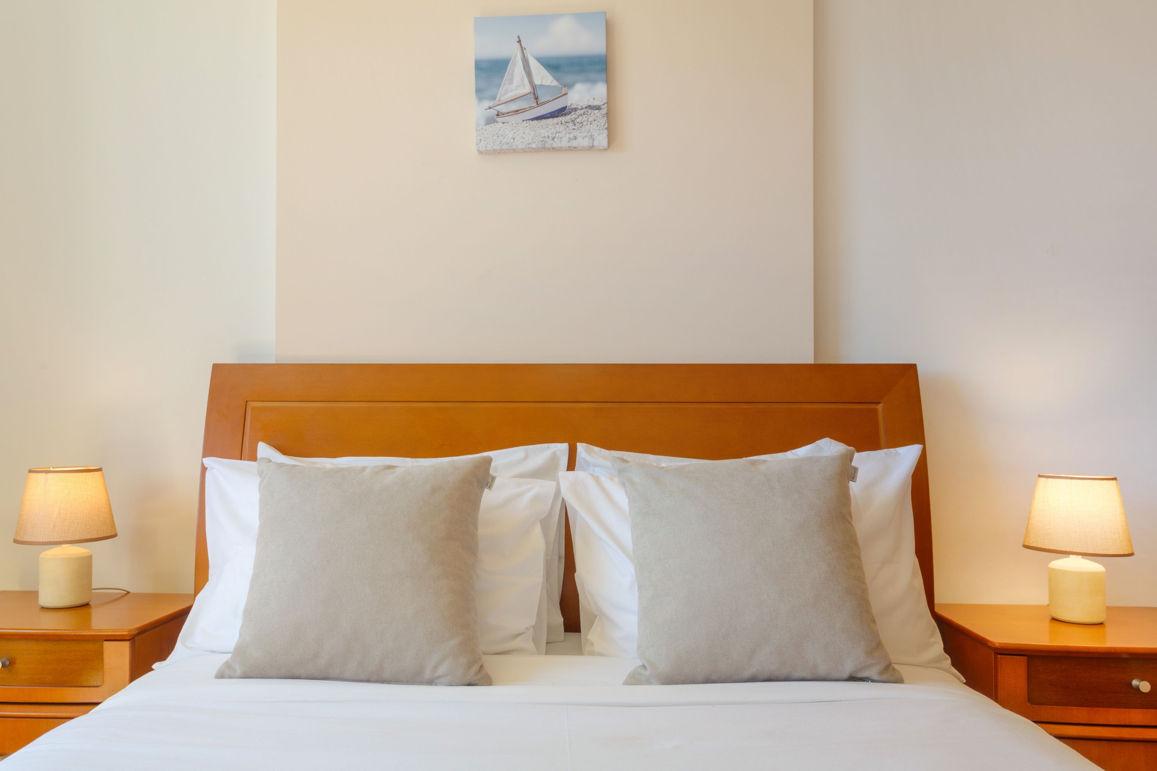 Habitación de hotel con cabecera de madera, dos almohadas blancas, un pequeño cuadro de un velero y dos lámparas de noche.