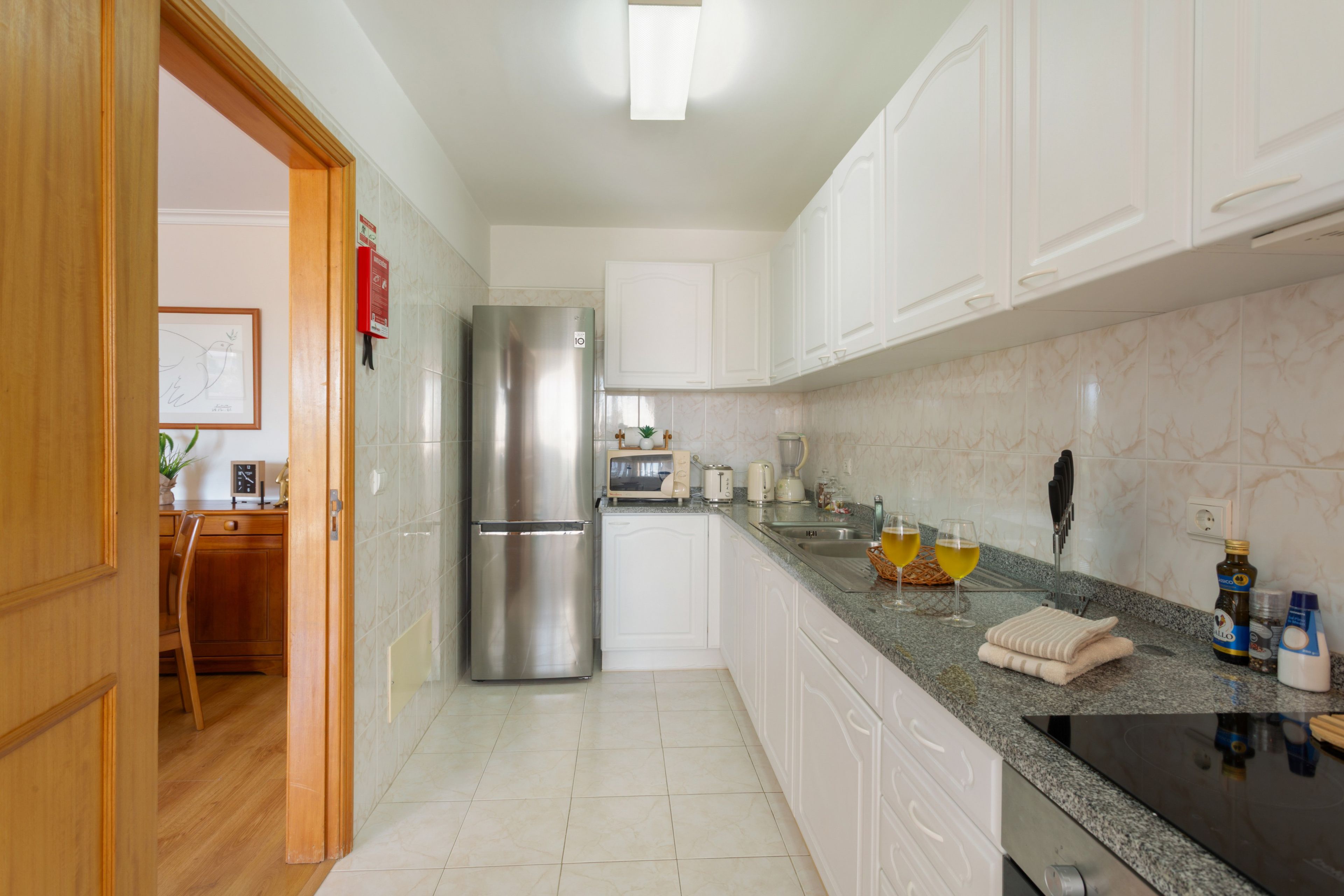 A modern kitchen with white cabinets, a stainless steel refrigerator, a granite countertop, and a dining area with a wooden table and chairs.