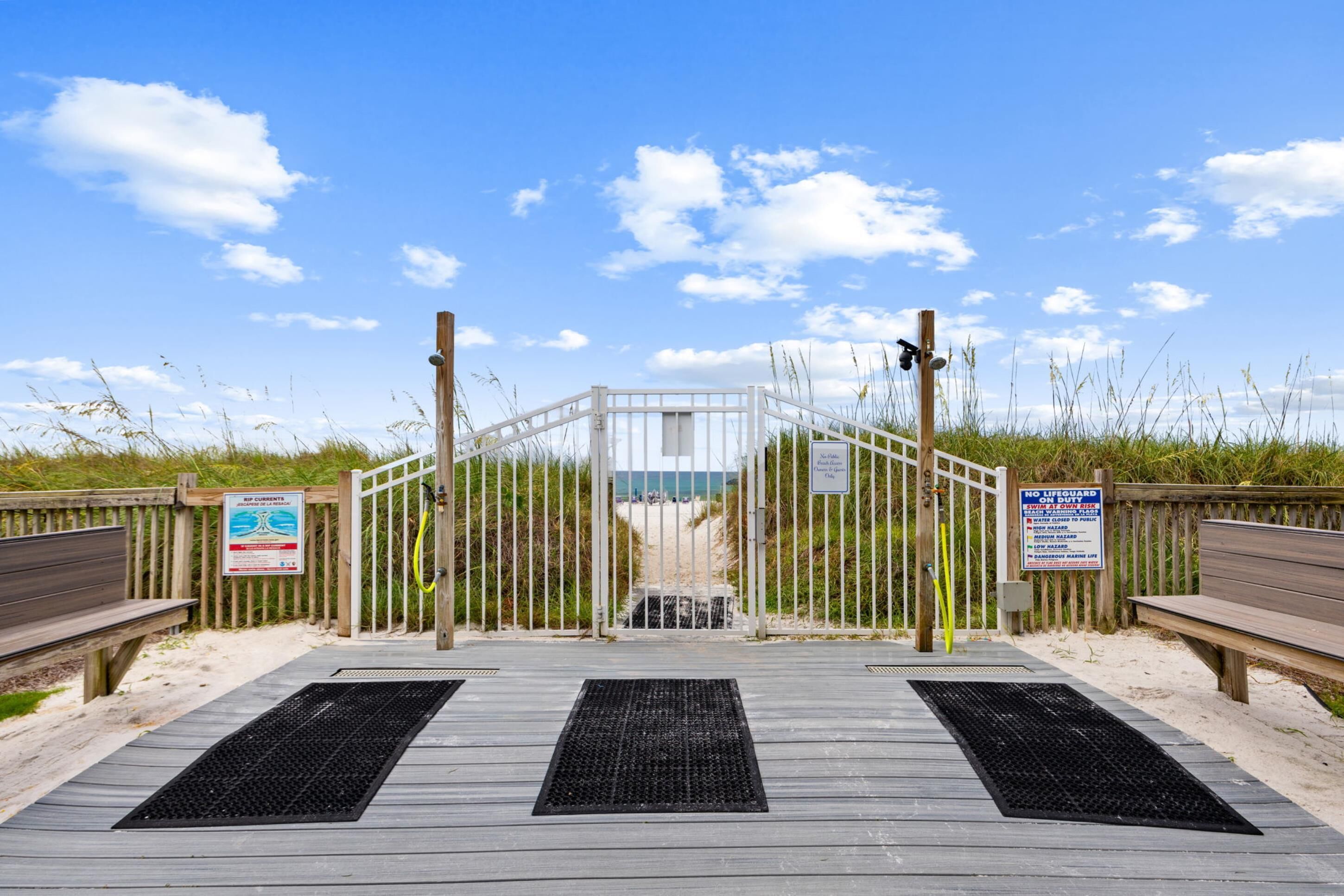 A beach entrance with a wooden boardwalk leading to a gated area, featuring warning signs and a lifeguard tower.