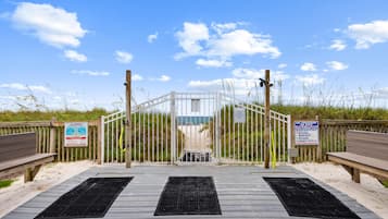 A beach entrance with a wooden boardwalk leading to a gated area, featuring warning signs and a lifeguard tower.