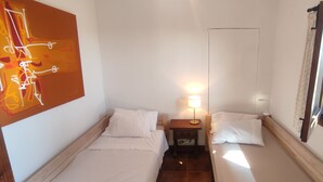 Ein kleiner, gut beleuchteter Raum mit zwei Betten, einem Nachttisch mit Lampe, einem Wandbild und einem Fenster mit Vorhängen.