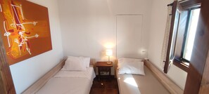 Ein kleiner, gut beleuchteter Raum mit zwei Betten, einem Nachttisch mit Lampe, einem Wandbild und einem Fenster mit Vorhängen.