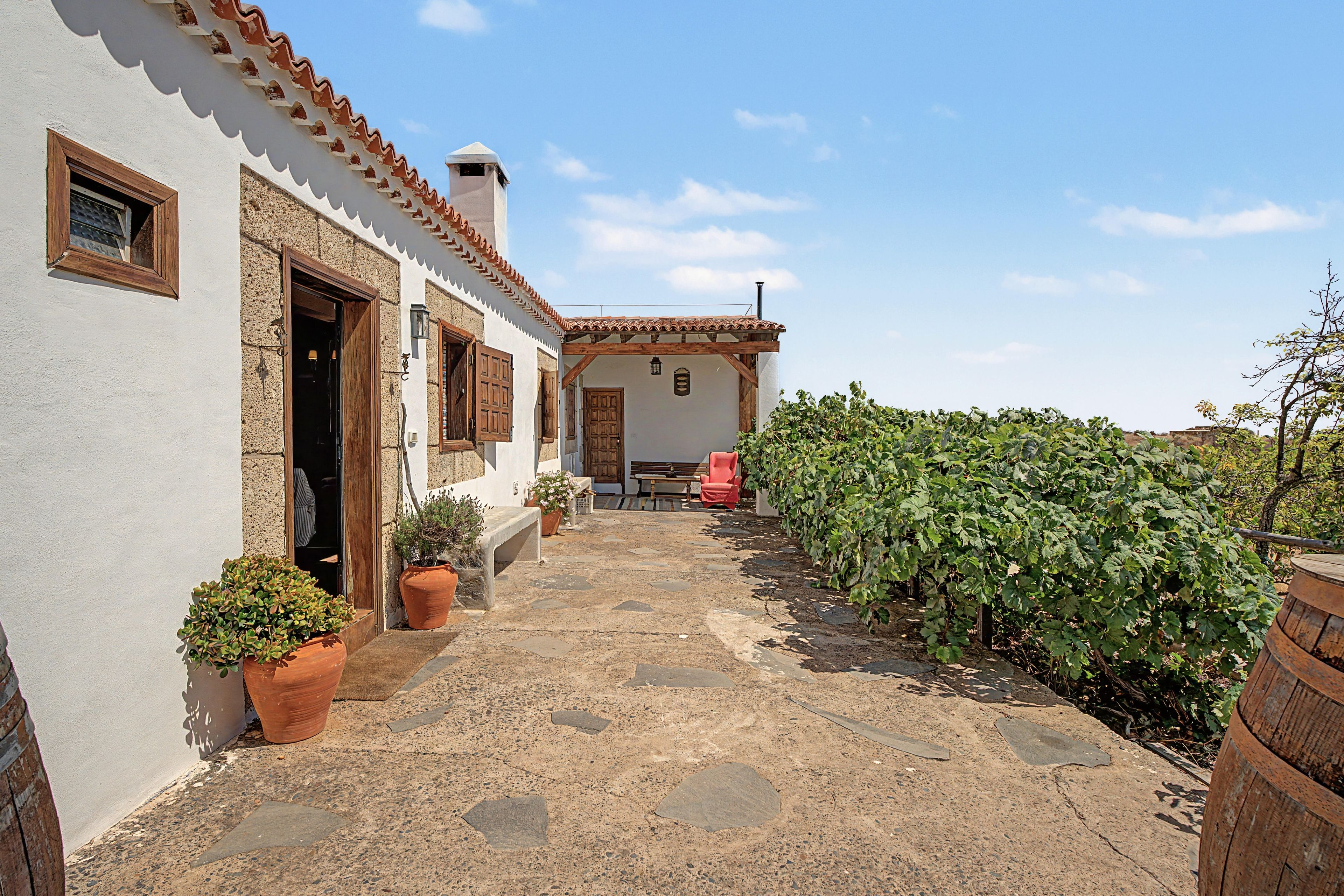 A courtyard with a stone pathway leading to a building with a tiled roof and a chimney. There are potted plants and a bench along the path.