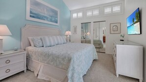 Une chambre à coucher comprenant un lit, des tables de chevet, une commode, un ventilateur de plafond, un tableau, un téléviseur et un placard avec un miroir.