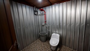 A compact bathroom with a toilet, a bucket, and a shower head against a corrugated metal wall. - ECO VILLAGE MALAGUFUK (Sorong Regency)