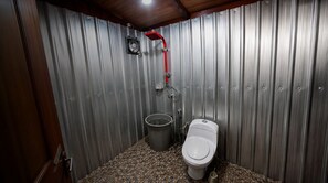 A compact bathroom with a toilet, a bucket, and a shower head against a corrugated metal wall. - ECO VILLAGE MALAGUFUK (Sorong Regency)
