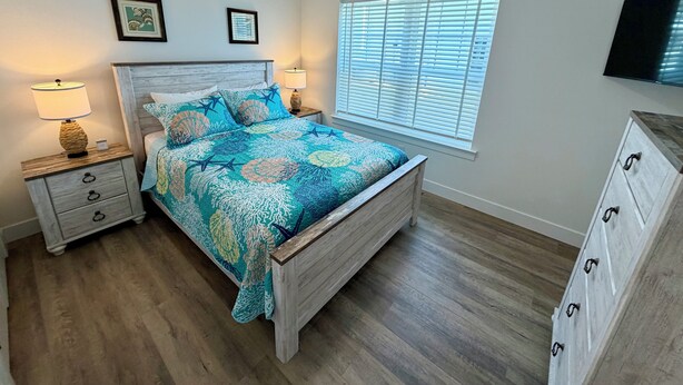Un dormitorio con cama, dos mesitas de noche, cómoda, ventana con persianas y ventilador de techo.
