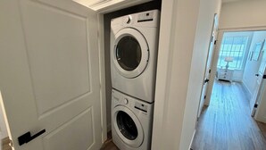 Un lavadero con dos lavadoras de carga frontal apiladas, una puerta que da a otra habitación y un pasillo con ventana y lámpara.