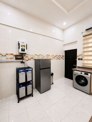 A compact kitchen with a refrigerator, washing machine, and a utility cart with batteries. - The Amber Stay | 24/7 Power| Fully Serviced |Lekki (Eti-Osa)