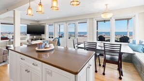 A spacious living area with a large island, dining table, and chairs, overlooking a beach view through large windows. - Hamptons Best Beach House. On the Sand. Sleeps 8. Parking. Porch. Beach Gear (Hampton)