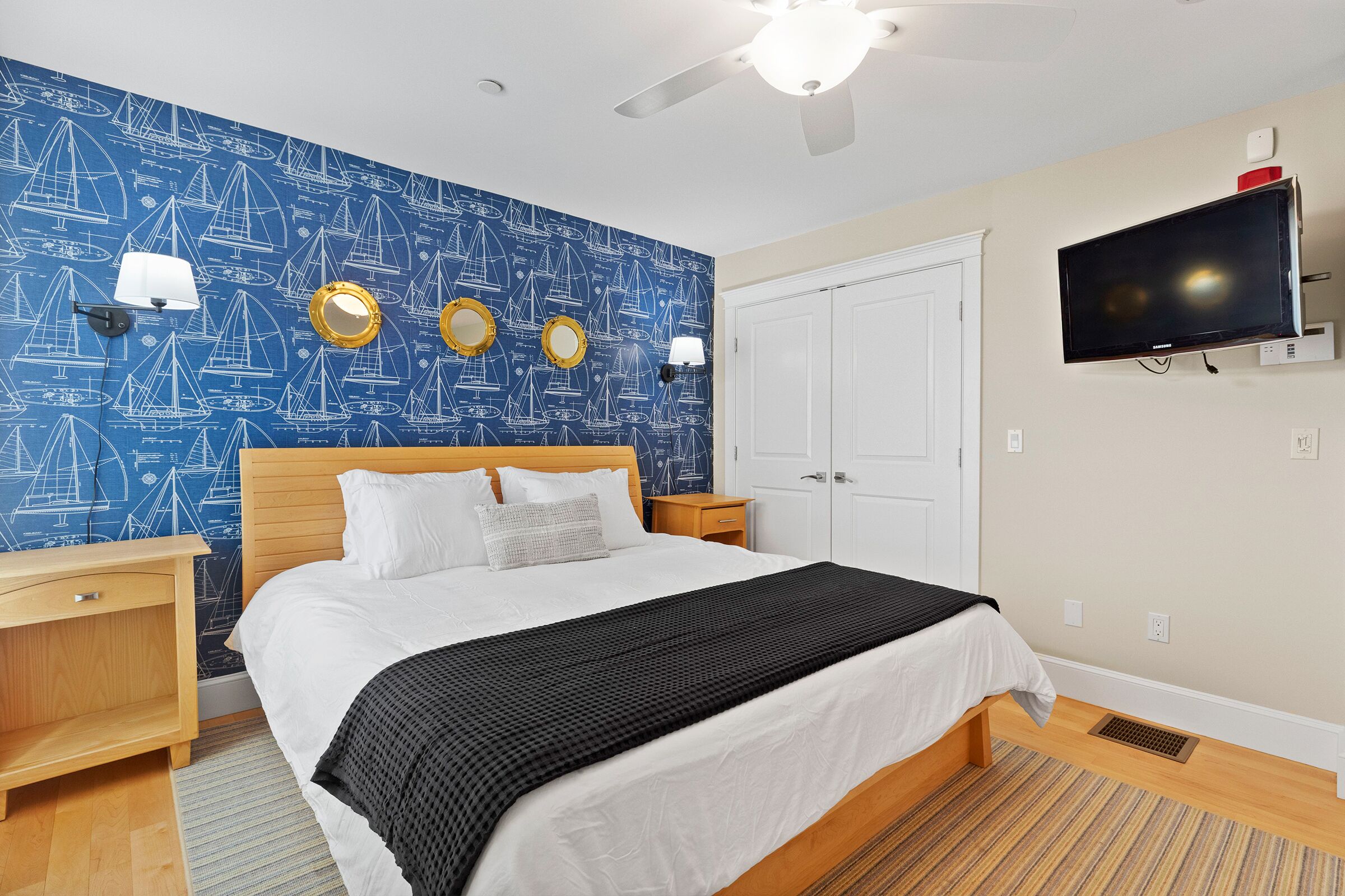 A hotel room with a bed, a TV, a ceiling fan, and a wall with nautical-themed wallpaper.
