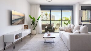 A modern living room with a sofa, a coffee table, a TV, and a large window with a view of buildings. - Elegant 4BR Stay l Balcony & Mamad (Bat Yam)
