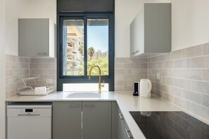 A modern kitchen with a built-in dishwasher, a window with a view of buildings and trees, and a coffee maker on the counter. - Elegant 4BR Stay l Balcony & Mamad (Rishon LeTsiyon)