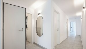 A modern hallway with white walls, a large mirror, and a door leading to a bathroom. - Elegant 4BR Stay l Balcony & Mamad (Bat Yam)