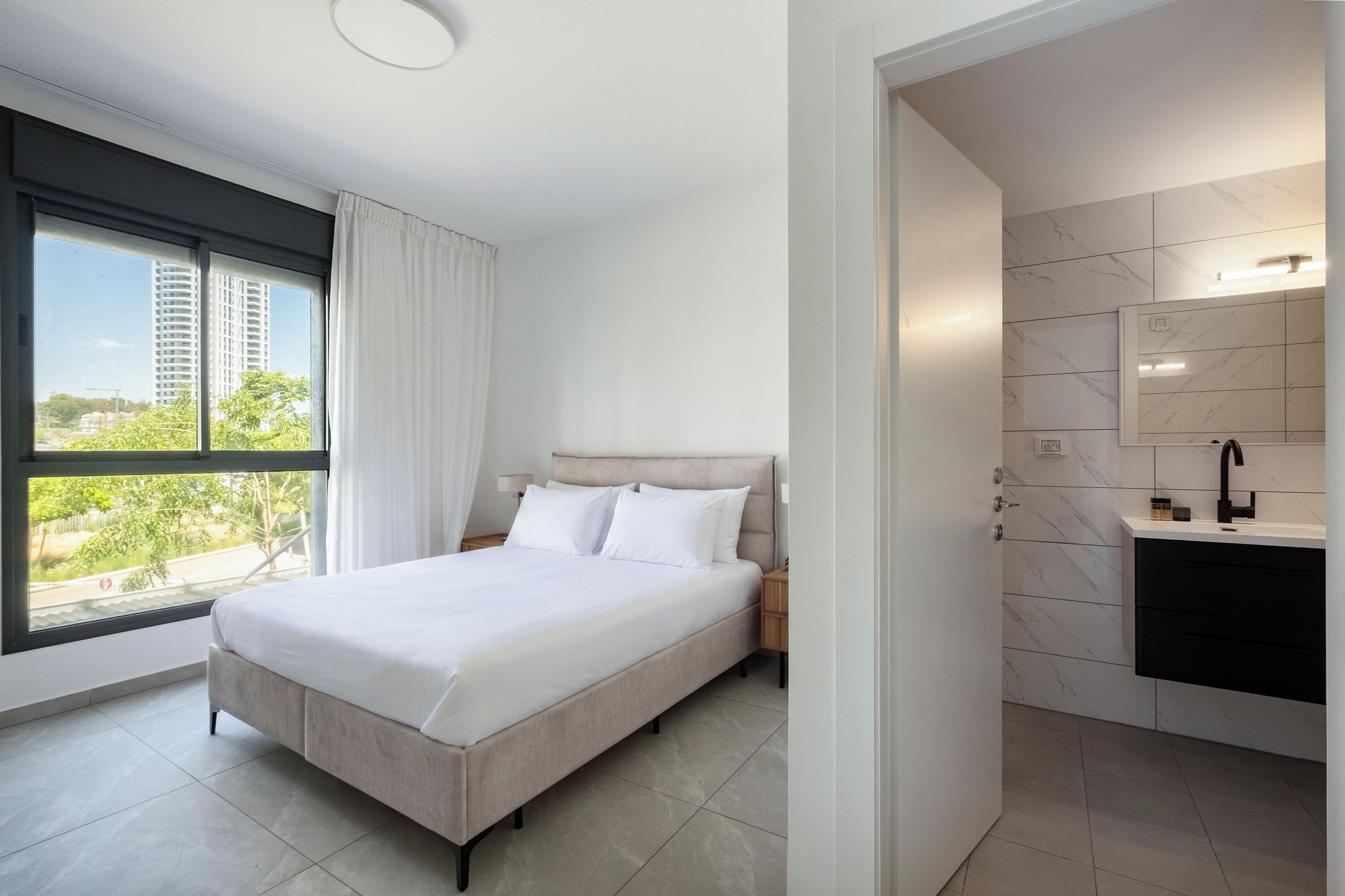 Habitación de hotel moderna con una cama grande, vistas a zonas verdes y edificios, baño con pared de mármol y una ventana con cortinas.