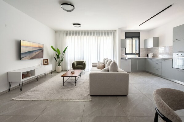 A modern living room with a sofa, a coffee table, and a TV on the wall. - Elegant 4BR Stay l Balcony & Mamad (Rishon LeTsiyon)