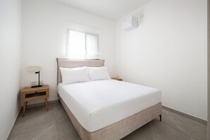 A minimalist bedroom with a bed, bedside table, lamp, and a window. - Elegant 4BR Stay l Balcony & Mamad (Rishon LeTsiyon)