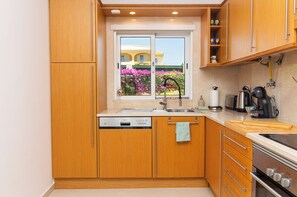 Eine Küche mit Holzausstattung, einem Geschirrspüler, einer Spüle, einer Kaffeemaschine und einem Fenster mit Blick auf ein Haus und Blumen.