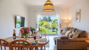 Ein Wohnzimmer mit einer Couch, einem Esstisch mit Stühlen, einem Fernseher und Blick auf einen Garten durch eine Schiebetür.