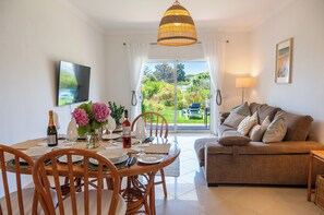 A living room with a sofa, dining table with chairs, a TV, and a view of a garden through a sliding door. - Casa Coelha - Garden Apartment near the Coast (Albufeira)