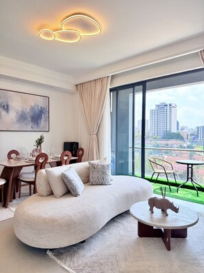 A modern living room with a curved sofa, dining area, and a view of a cityscape through large windows. - The Velvet Nook Rooftop Pool and Gym (Nairobi)