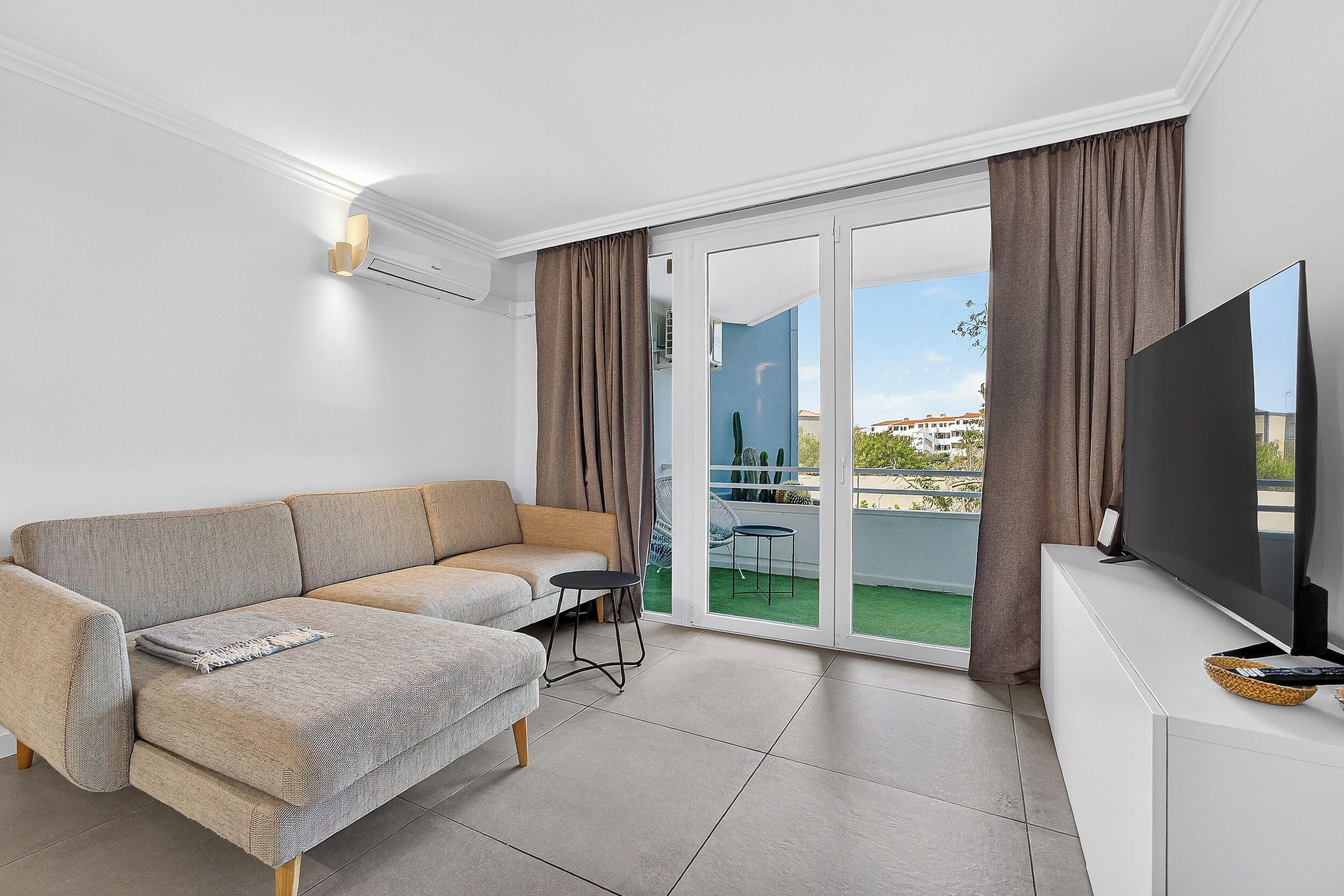 A modern living room with a sofa, a small table, a flat-screen TV, and a sliding glass door leading to a balcony.