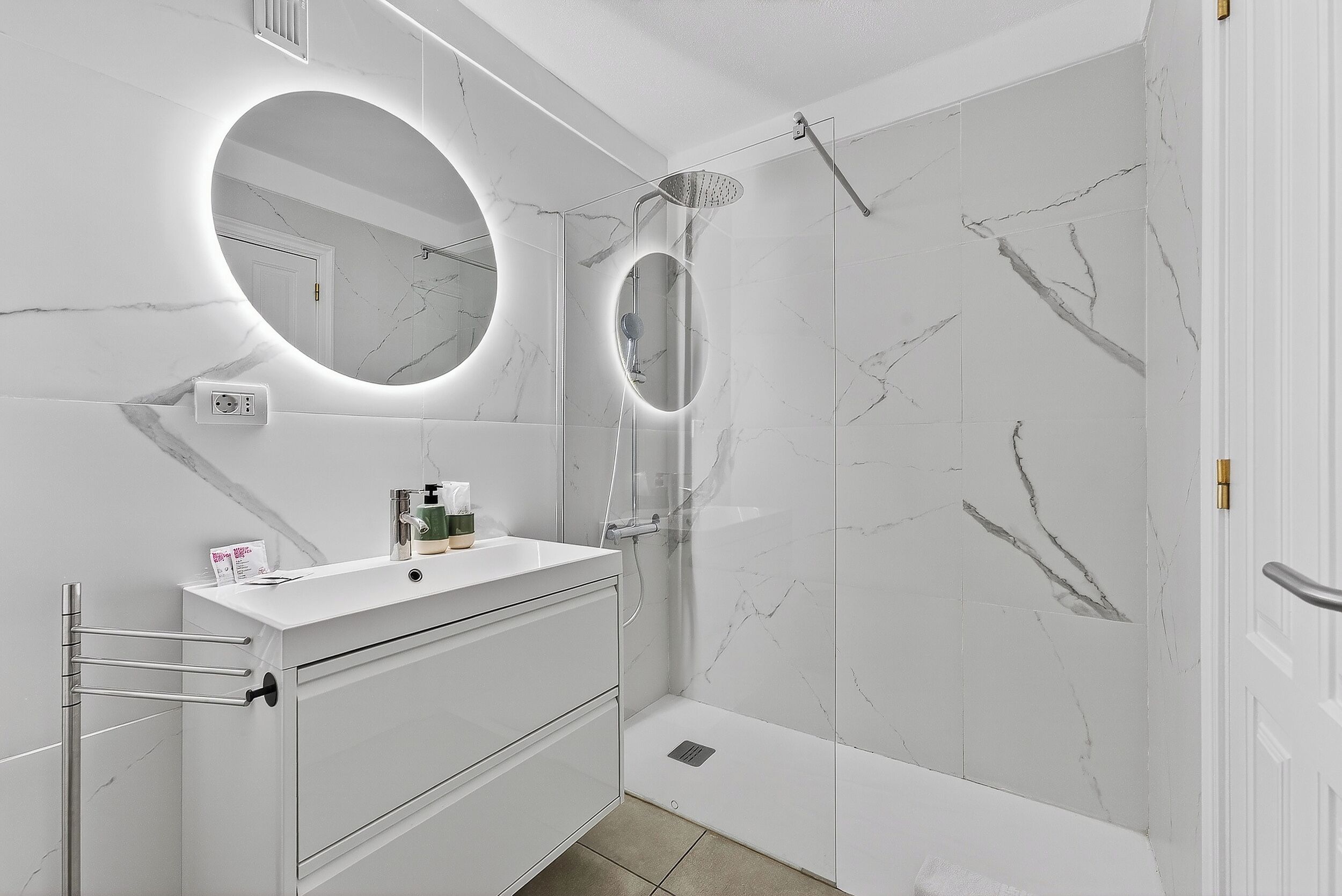 Een moderne badkamer met een marmeren wand, een ronde spiegel, een douche en een witte wastafelmeubel.