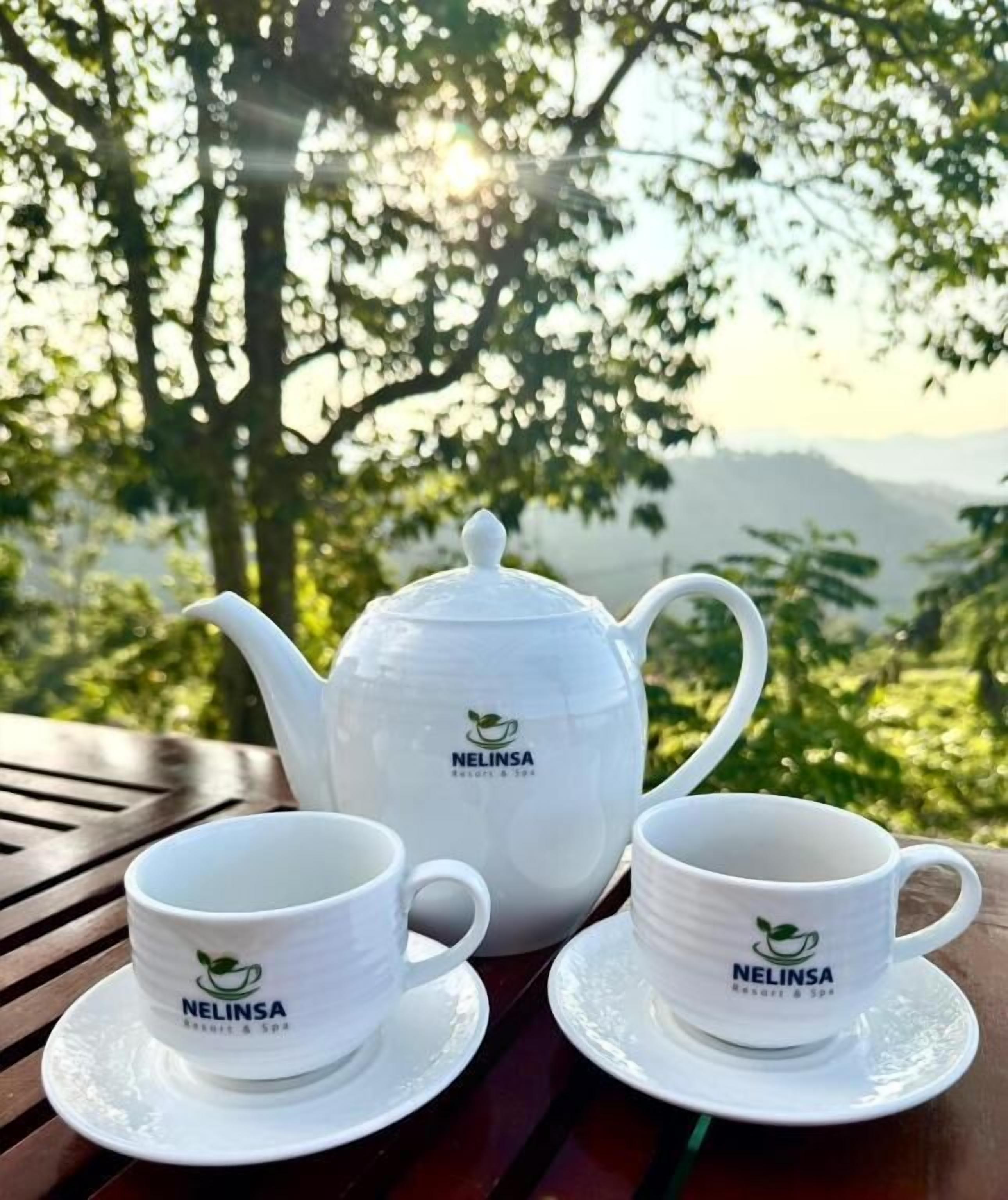 Un set da tè bianco con marchio 'NELINSA' su un tavolo di legno all'aperto, con vista su alberi e colline.