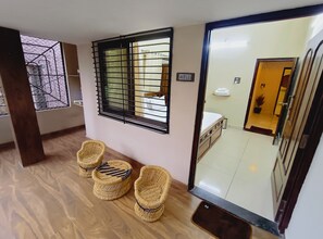 A hotel room with a large mirror, a window with metal bars, a bed, a bedside table, and two wicker chairs. - Mango Tree 3BHK Apartment (Ujjain)