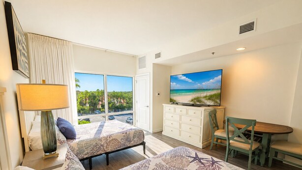 A hotel room with a bed, a desk, a television, and a view of the beach. - 4th-Floor Studio | Walk to Beach | Pool, Beach Tram | Luau II 6421, Sandestin (Miramar Beach)
