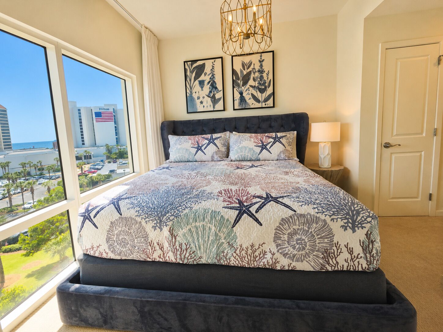 A hotel room with a large window, a bed with a patterned bedspread, a chandelier, and two framed botanical prints on the wall.
