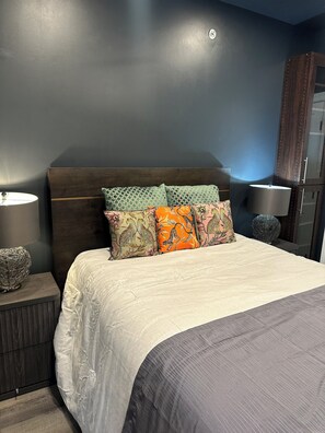 Ein Schlafzimmer mit dunklem Kopfteil, einem Bett mit gemusterten Kissen, einer Nachttischlampe und einem wandmontierten Spiegel.
